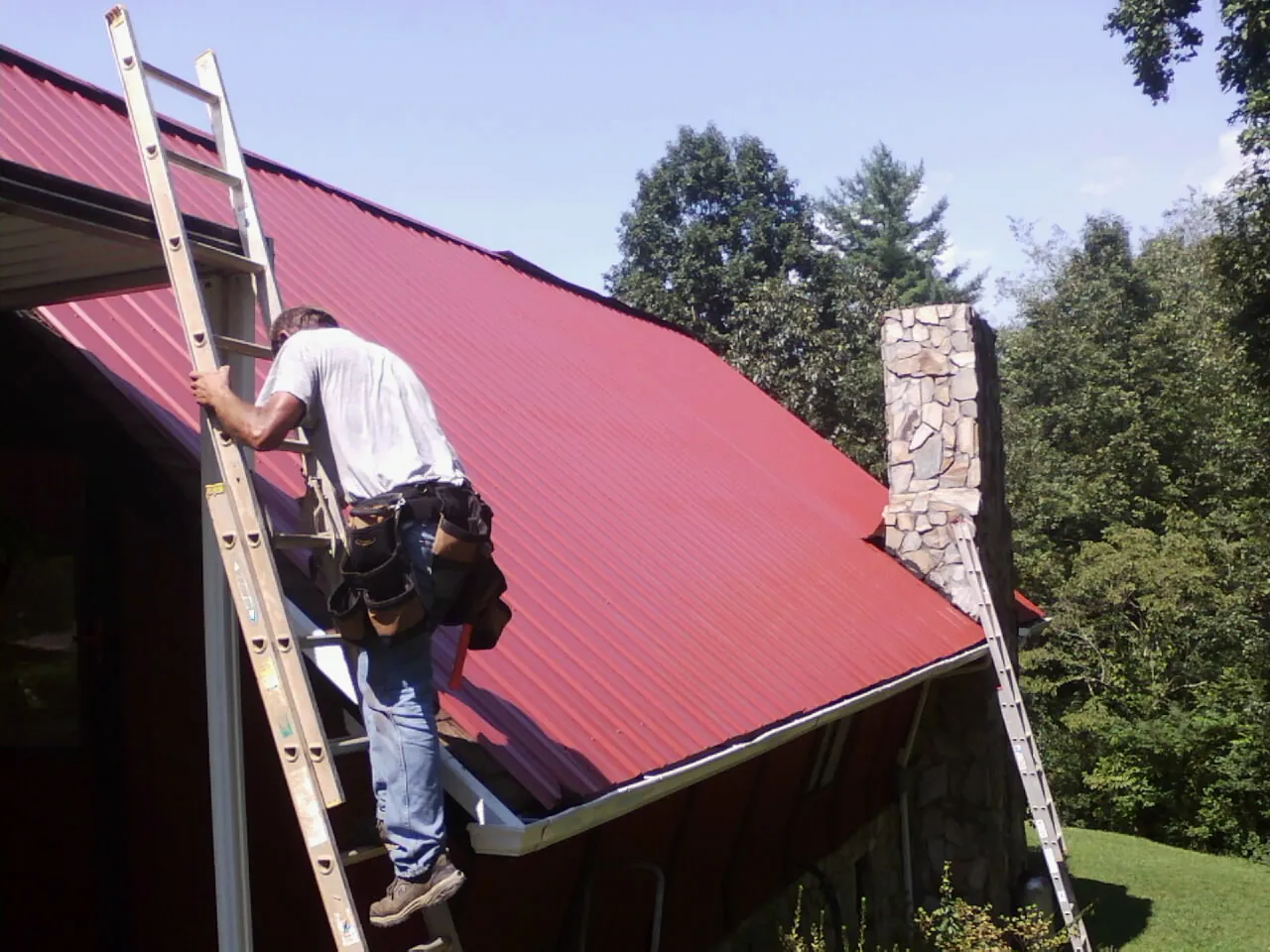 Roofer on a ladder beside a red metal roof and stone chimney — Soffit & Fascia Repair by Roofing Brothers in Brunsville